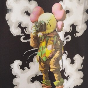 May include: Black t-shirt featuring the text "OFF WHITE" in white, with a cartoon astronaut design. The astronaut is holding a camera and is surrounded by white cloud-like shapes. The text "WE CAME IN PEACE FOR ALL MANKIND" is also visible.