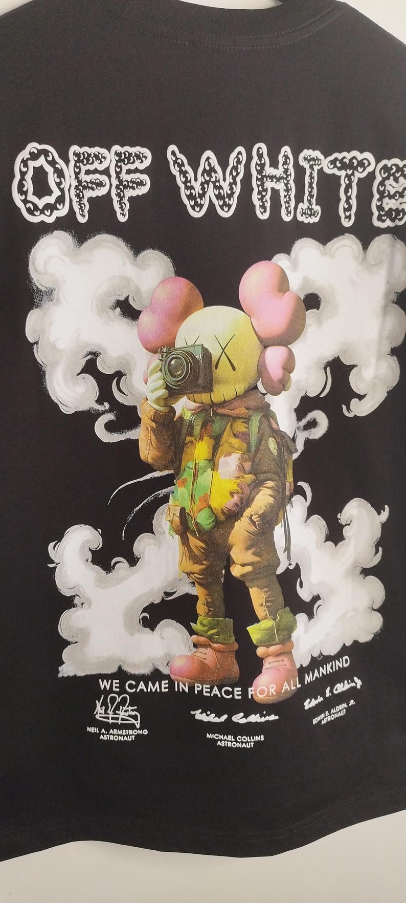 May include: Black t-shirt featuring the text "OFF WHITE" in white, with a cartoon astronaut design. The astronaut is holding a camera and is surrounded by white cloud-like shapes. The text "WE CAME IN PEACE FOR ALL MANKIND" is also visible.