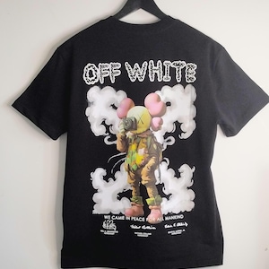 May include: Black t-shirt with the text "OFF WHITE" in white and a graphic of a cartoon figure holding a camera, surrounded by white clouds. The phrase "WE CAME IN PEACE FOR ALL MANKIND" is printed below the graphic, along with astronaut names.