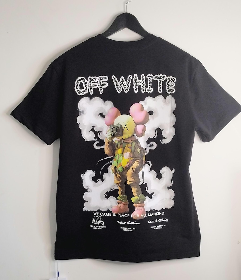 May include: Black t-shirt with the text "OFF WHITE" in white and a graphic of a cartoon figure holding a camera, surrounded by white clouds. The phrase "WE CAME IN PEACE FOR ALL MANKIND" is printed below the graphic, along with astronaut names.