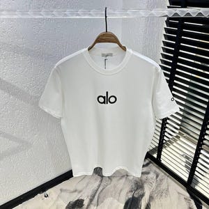 Puede incluir: Camiseta blanca de cuello redondo con la palabra "alo" impresa en negro en la parte delantera. La camiseta est&aacute; colgada de una percha de madera. La manga tiene un peque&ntilde;o logotipo. El fondo es una pared blanca texturizada.