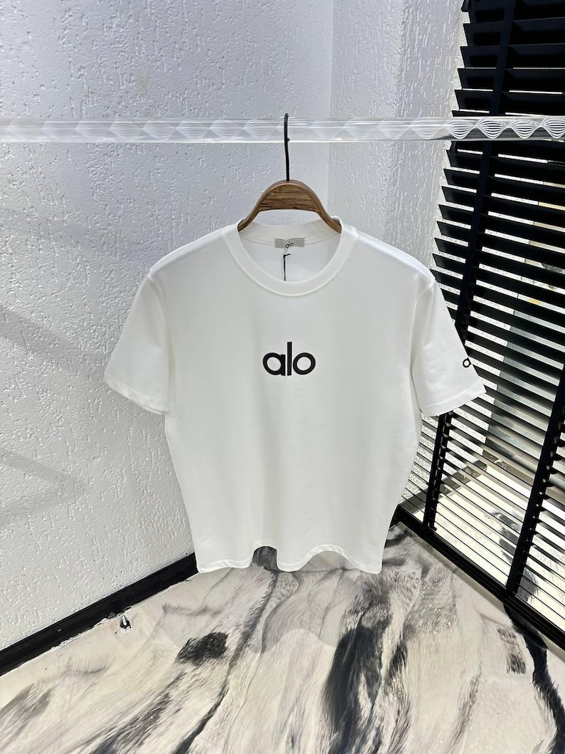 Puede incluir: Camiseta blanca de cuello redondo con la palabra "alo" impresa en negro en la parte delantera. La camiseta est&aacute; colgada de una percha de madera. La manga tiene un peque&ntilde;o logotipo. El fondo es una pared blanca texturizada.