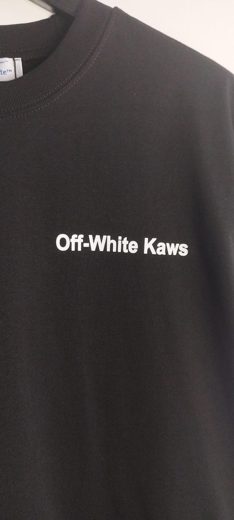 May include: Dark charcoal gray sweatshirt with the text "Off-White Kaws" in white block letters. The sweatshirt is made of a soft material and has a crew neck. The image is a close-up of the front of the shirt.