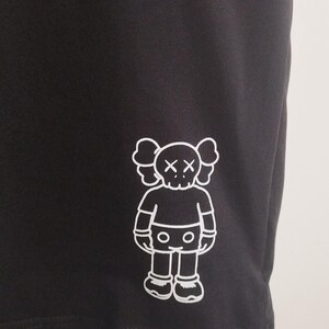 May include: Black athletic shorts with a white graphic of a cartoon figure with crossed-out eyes. The figure has a rounded head, arms, legs, and feet. The shorts are made of a smooth, dark fabric.