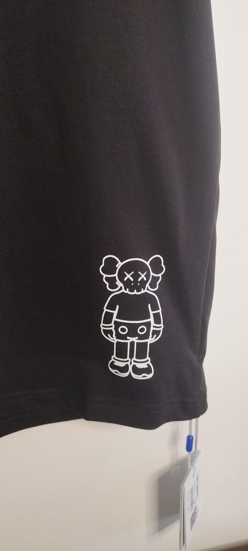 May include: Black athletic shorts with a white graphic of a cartoon figure with crossed-out eyes. The figure has a rounded head, arms, legs, and feet. The shorts are made of a smooth, dark fabric.