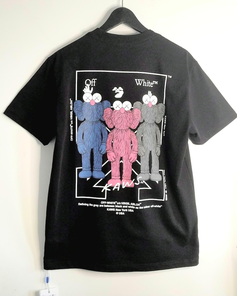 May include: Black t-shirt featuring three cartoon figures in blue, pink, and gray, with the text "OFF-WHITE" and "KAWS" in white. The figures have X-shaped eyes and are standing in a row. The shirt is hanging on a black hanger.
