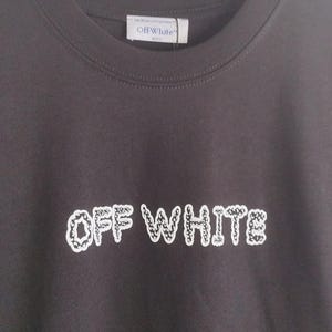 May include: Dark gray t-shirt with the text "OFF WHITE" in a white, patterned font. The shirt is hanging on a black hanger. The brand label "Off White" is visible at the neckline.