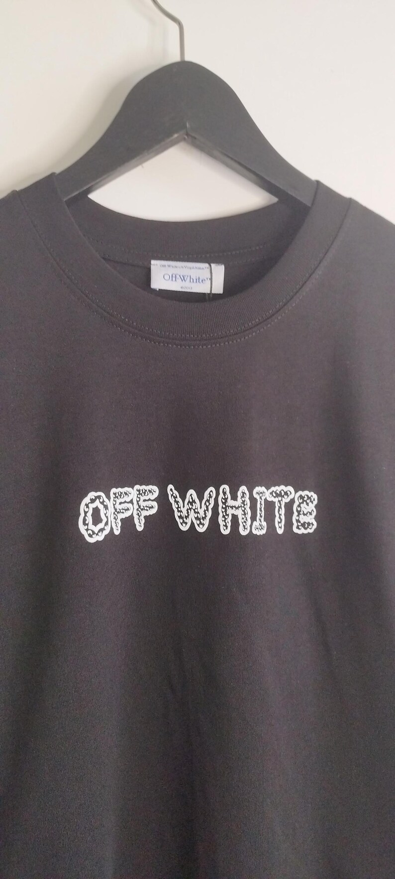 May include: Dark gray t-shirt with the text "OFF WHITE" in a white, patterned font. The shirt is hanging on a black hanger. The brand label "Off White" is visible at the neckline.
