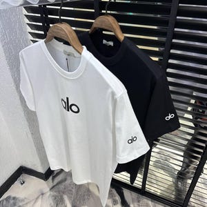 Puede incluir: Dos camisetas de manga corta, una blanca y otra negra, colgadas de perchas de madera. La camiseta blanca tiene "alo" impreso en negro en la parte delantera y en la manga izquierda. La camiseta negra tiene "alo" en la manga izquierda.
