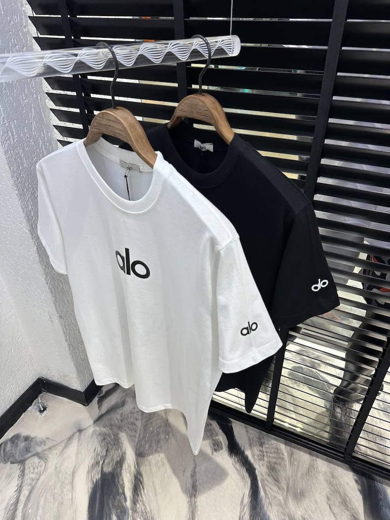 Puede incluir: Dos camisetas de manga corta, una blanca y otra negra, colgadas de perchas de madera. La camiseta blanca tiene "alo" impreso en negro en la parte delantera y en la manga izquierda. La camiseta negra tiene "alo" en la manga izquierda.
