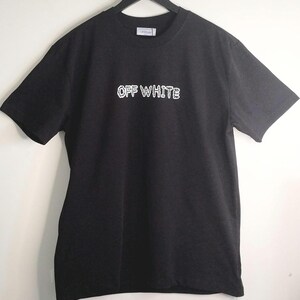 May include: Black t-shirt with the text "OFF WHITE" in white, displayed on a black hanger. The shirt has short sleeves and a crew neck. The text appears to be a brand name.