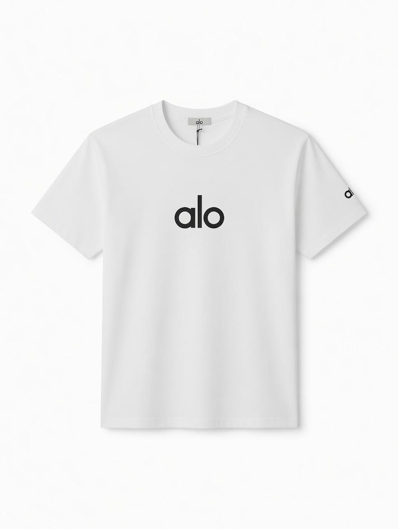 Puede incluir: Camiseta blanca de cuello redondo con la palabra "alo" impresa en negro en el pecho. El logotipo de la marca tambi&eacute;n est&aacute; en la manga. La camiseta tiene un dise&ntilde;o simple y minimalista.