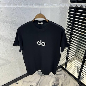 Op de afbeelding: Zwart T-shirt met ronde hals en de witte tekst "alo" gecentreerd op de voorkant. Het shirt hangt aan een houten hanger. Het shirt heeft korte mouwen en een label op de achterkant van de nek.