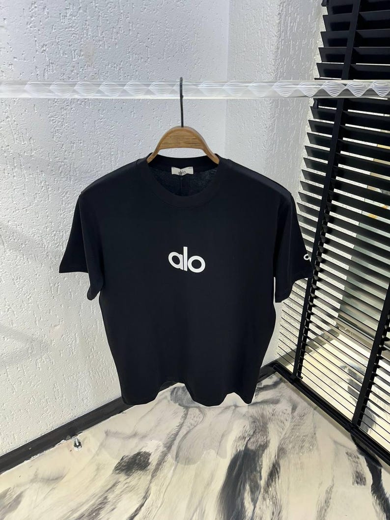 Op de afbeelding: Zwart T-shirt met ronde hals en de witte tekst "alo" gecentreerd op de voorkant. Het shirt hangt aan een houten hanger. Het shirt heeft korte mouwen en een label op de achterkant van de nek.