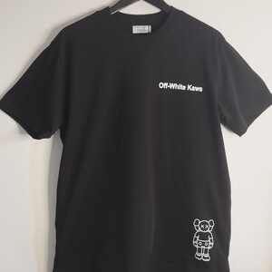 May include: Black t-shirt with the text "Off-White Kaws" in white on the front. A white cartoon figure is on the lower right side. The shirt is hanging on a black hanger.