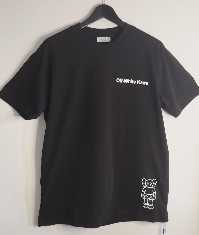 May include: Black t-shirt with the text "Off-White Kaws" in white on the front. A white cartoon figure is on the lower right side. The shirt is hanging on a black hanger.