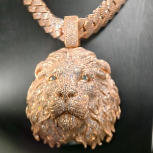 Peut inclure: Un collier avec un pendentif en forme de tête de lion couleur or rose, doté d'une chaîne épaisse à maillons cubains texturés. Le visage du lion est entièrement incrusté de nombreuses petites pierres claires et réfléchissantes scintillantes.