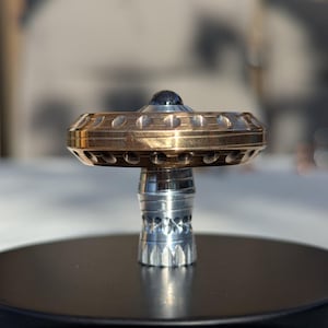 Peut inclure: Gros plan détaillé d'une toupie en métal. La toupie présente un anneau extérieur de couleur or avec des trous espacés, une base argentée et une sphère centrale sombre, peut-être noire. La toupie est posée sur une base circulaire noire.