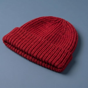 Può includere: Un berretto in maglia rosso scuro caratterizzato da una spessa trama a coste e un ampio risvolto ripiegato. Il cappello è realizzato con un motivo a maglia verticale coerente che corre dalla corona fino al bordo, creando un aspetto strutturato.