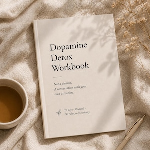 May include: A hardcover book titled "Dopamine Detox Workbook" with the text "Not a cleanse. A conversation with your own attention. 28 days. Undated. No rules, only curiosity." A cup of coffee and a pen are also in the image.