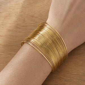 Può includere: Un ampio bracciale a fascia composto da numerosi sottili cerchi in filo metallico color oro sovrapposti per creare un aspetto stratificato e testurizzato che avvolge il polso con una finitura metallica uniforme e continua.