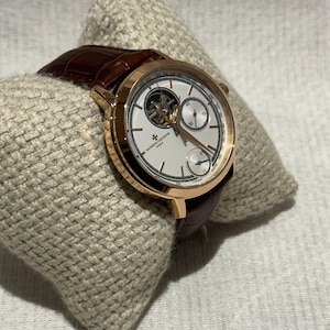 May include: A high-end wristwatch with a brown leather strap and a gold-toned case. The watch face is white with black and gold detailing, including subdials and complex mechanical elements. The watch is displayed on a textured, light-coloured surface.