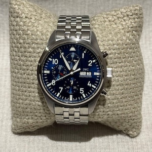 Könnte beinhalten: Eine Edelstahl-Armbanduhr mit blauem Zifferblatt, weißen Ziffern und Zeigern. Die Uhr verfügt über eine Chronographenfunktion und eine Datumsanzeige. Die Uhr wird auf einem beigen Kissen präsentiert. Das Zifferblatt zeigt "IWC SCHAFFHAUSEN".