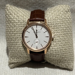 Puede incluir: Un reloj de pulsera dorado con esfera blanca y correa de cuero marrón. El reloj tiene un pequeño dial de segundos y se muestra sobre un cojín beige. La esfera del reloj presenta agujas e indicadores horarios negros.