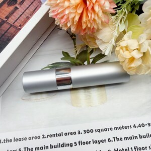 May include: A cylindrical silver metal spray bottle with a polished chrome center band. The container features a smooth matte finish and a sleek design, positioned horizontally against a backdrop of peach and cream-colored artificial flowers.