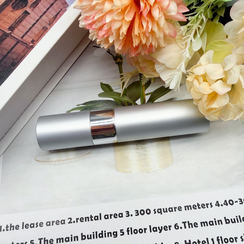 May include: A cylindrical silver metal spray bottle with a polished chrome center band. The container features a smooth matte finish and a sleek design, positioned horizontally against a backdrop of peach and cream-colored artificial flowers.