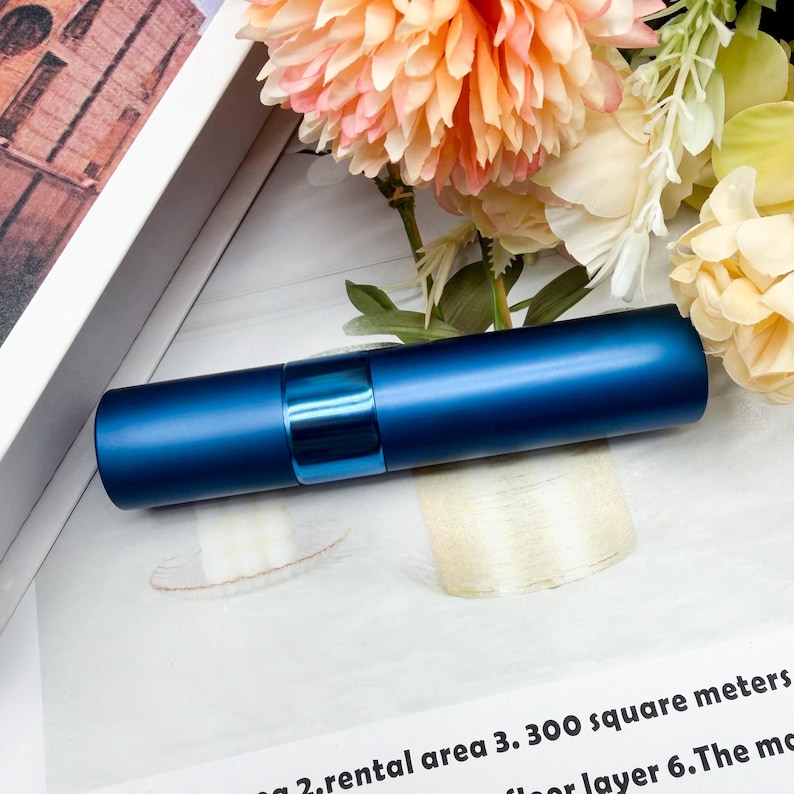 May include: A cylindrical, matte navy blue perfume atomizer with a glossy, metallic blue center band. The container features a sleek, minimalist design with a smooth finish, positioned horizontally against a backdrop of soft, light-colored flowers.