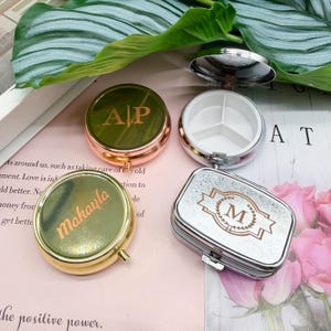 May include: Four personalized pill cases in various shapes and colors. One round case is open, revealing three compartments. The cases are gold, silver, and rose gold, with initials and a name engraved on them.
