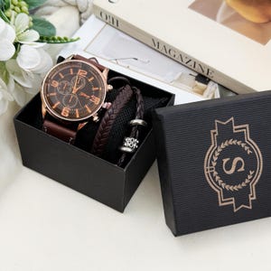 Op de afbeelding: Een roségoudkleurig polshorloge met een bruine wijzerplaat en bruine leren band, gecombineerd met een donkerbruine gevlochten leren armband met zilverkleurige metalen kralen, gepresenteerd in een zwarte geschenkdoos met een gouden S-logo.