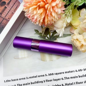 May include: A cylindrical purple atomizer bottle with a matte finish and a glossy metallic purple band around the center. The container is a solid, opaque purple color and features a smooth, uniform surface design for holding liquid contents.