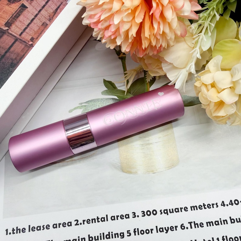May include: A cylindrical, matte mauve lipstick tube with a metallic silver band and a small heart icon printed above the word "CONNER" in white lettering. The tube is positioned horizontally against a background of light peach flowers.