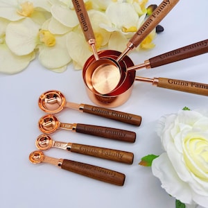 May include: A set of copper measuring cups and spoons with dark wood handles. Each handle is engraved with the text Guina Family. The cups are nested together, and the spoons are arranged in a row, featuring measurements like 1 TSP and 1/2 TSP.