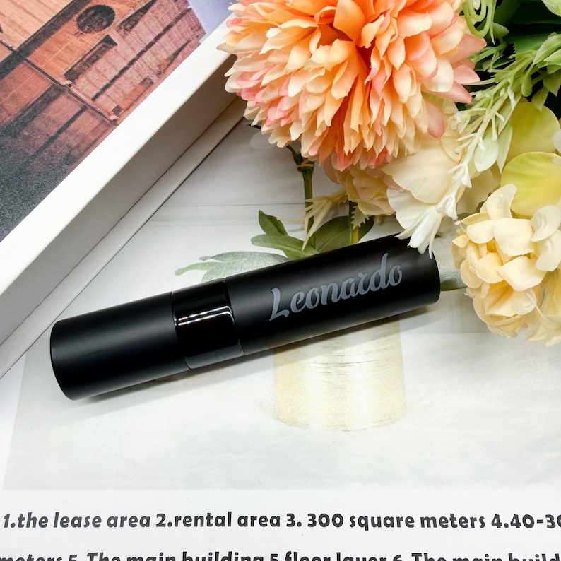 May include: A matte black cylindrical tube featuring the name Leonardo engraved in a cursive script. The tube has a glossy black band near the center and is positioned horizontally against a background of peach-colored flowers and text.
