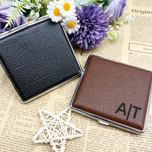 May include: Two square cases with silver metal frames and textured surfaces. One case is black, and the other is brown with the black letters A|T printed on the front. Both items feature a hinged closure and a slim, compact, flat design.