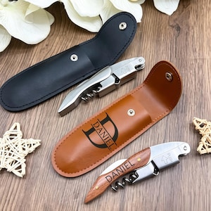 May include: Two waiter-style corkscrews with folding metal mechanisms and integrated foil cutters. One corkscrew has a wood-grain handle engraved with the name DANIEL, paired with a tan case featuring the name DANIEL. The other is plain metal with a black case.