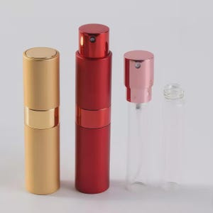 May include: Four cylindrical perfume atomizers are displayed. One is solid gold, one is solid red, one has a metallic pink spray nozzle on a clear glass vial, and one is a clear glass vial with a threaded neck and no cap attached.