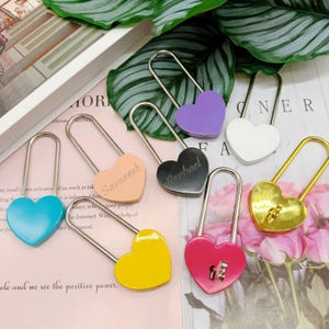 Peut inclure: Plusieurs cadenas en forme de cœur de différentes couleurs : bleu, pêche, noir, blanc, jaune, or et rouge. Certains cadenas sont gravés de noms. Les cadenas ont des anses en métal argenté.