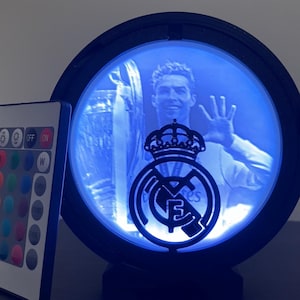 Puede incluir: Una l&aacute;mpara LED redonda con marco negro, que muestra a un jugador de f&uacute;tbol y el logotipo de un equipo. La luz emite un brillo azul, y se ve un mando a distancia con opciones de color. El logotipo del equipo est&aacute; en el centro.