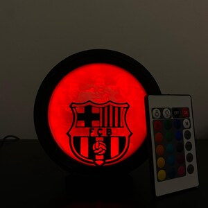 Puede incluir: L&aacute;mpara LED redonda con el escudo del FC Barcelona iluminado en rojo dentro de un marco negro. Un mando a distancia con opciones de color est&aacute; a la derecha. La l&aacute;mpara est&aacute; sobre una superficie oscura.