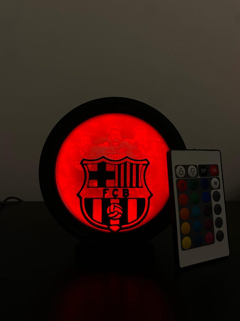 Puede incluir: L&aacute;mpara LED redonda con el escudo del FC Barcelona iluminado en rojo dentro de un marco negro. Un mando a distancia con opciones de color est&aacute; a la derecha. La l&aacute;mpara est&aacute; sobre una superficie oscura.