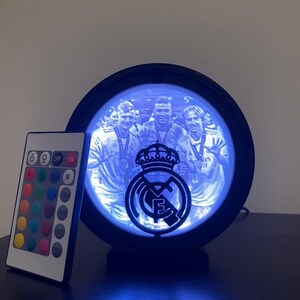 Puede incluir: L&aacute;mpara LED redonda negra con el escudo del Real Madrid y una foto del equipo. La l&aacute;mpara emite un brillo azul e incluye un mando a distancia con opciones de color. Sobre una superficie oscura.