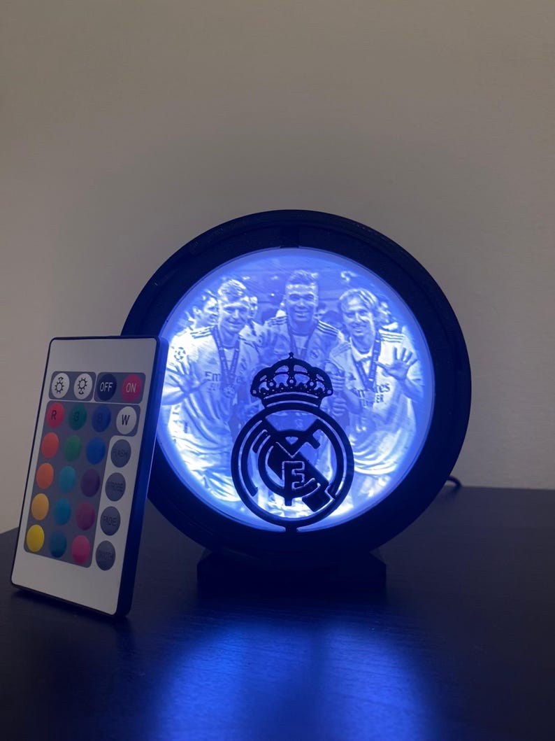 Puede incluir: L&aacute;mpara LED redonda negra con el escudo del Real Madrid y una foto del equipo. La l&aacute;mpara emite un brillo azul e incluye un mando a distancia con opciones de color. Sobre una superficie oscura.