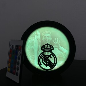Puede incluir: L&aacute;mpara LED redonda con marco negro, que muestra una imagen te&ntilde;ida de verde de un futbolista y el logotipo del Real Madrid. Un mando a distancia con opciones de color est&aacute; a la izquierda. La l&aacute;mpara est&aacute; sobre una superficie oscura.