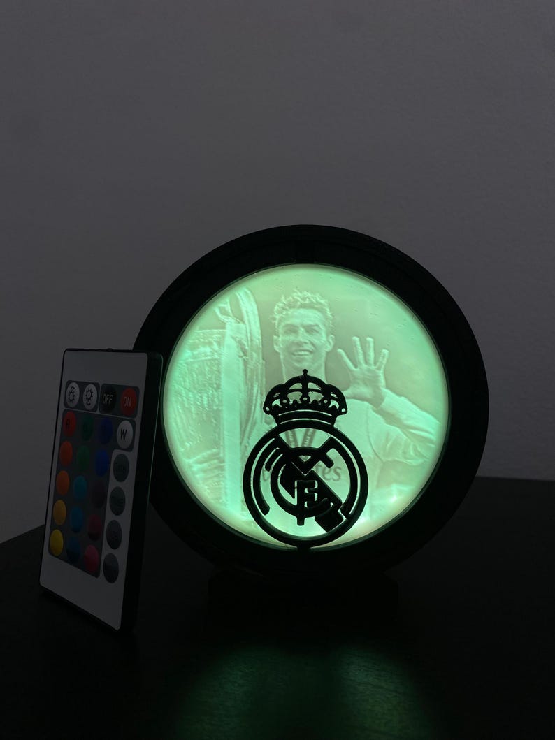 Puede incluir: L&aacute;mpara LED redonda con marco negro, que muestra una imagen te&ntilde;ida de verde de un futbolista y el logotipo del Real Madrid. Un mando a distancia con opciones de color est&aacute; a la izquierda. La l&aacute;mpara est&aacute; sobre una superficie oscura.
