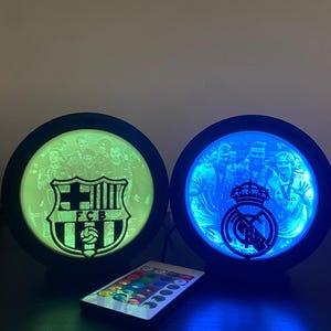 Puede incluir: Dos l&aacute;mparas redondas iluminadas con logotipos de equipos de f&uacute;tbol, una verde y otra azul. La l&aacute;mpara izquierda muestra el logotipo del FCB, mientras que la derecha muestra el logotipo del Real Madrid. Un mando a distancia con opciones de color est&aacute; debajo.