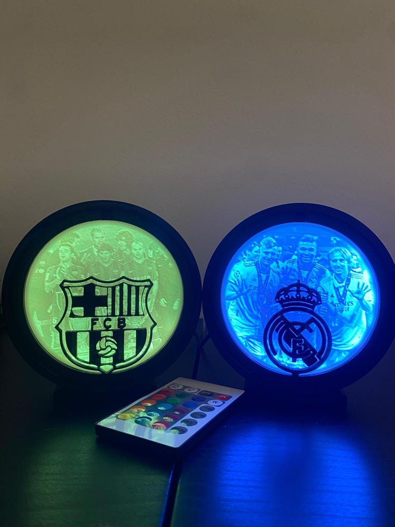 Puede incluir: Dos l&aacute;mparas redondas iluminadas con logotipos de equipos de f&uacute;tbol, una verde y otra azul. La l&aacute;mpara izquierda muestra el logotipo del FCB, mientras que la derecha muestra el logotipo del Real Madrid. Un mando a distancia con opciones de color est&aacute; debajo.